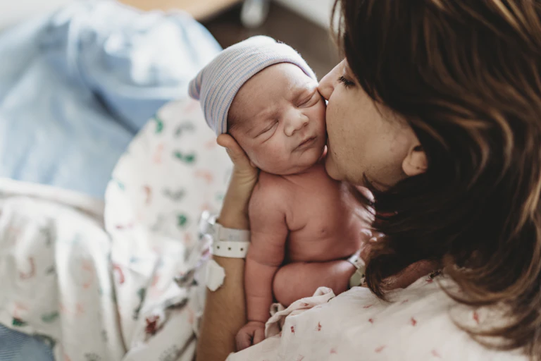Woman in hospital gown kisses her tiny newborn's cheek.