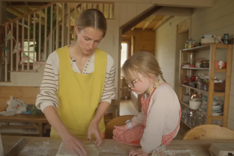 A woman wearing an apron rolls out dough while a young girl watches