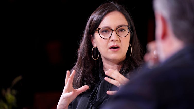 A woman wearing glasses speaks to a man on stage.
