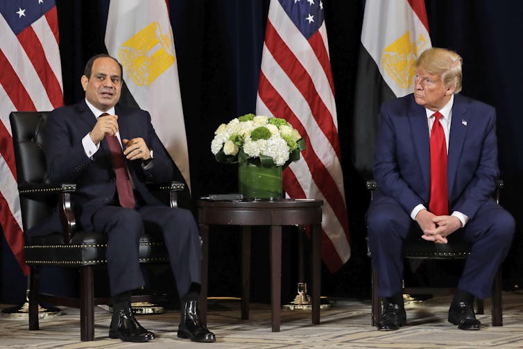 Two men seated chat surrounded by flags.