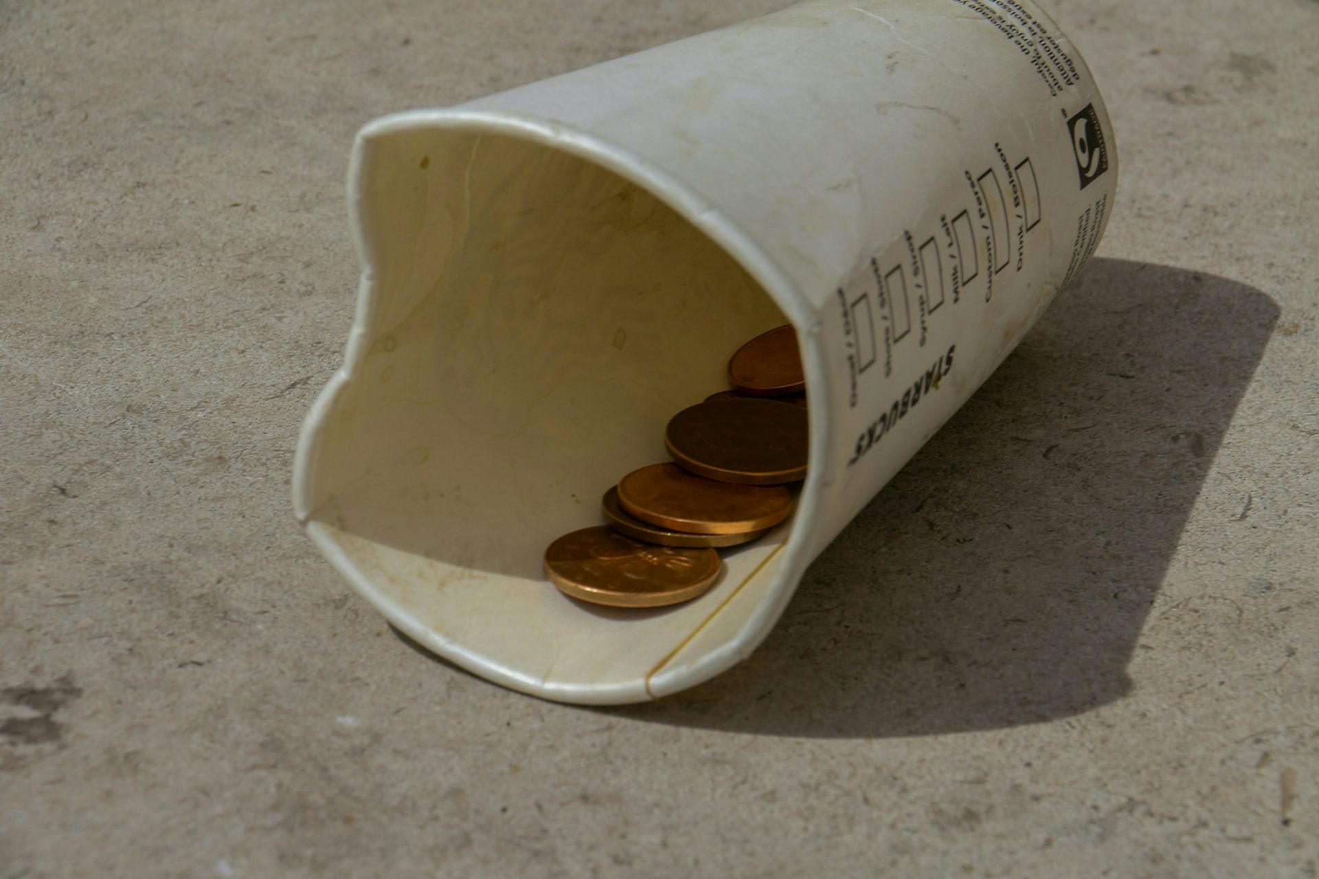 An old and empty Starbucks coffee cup lays on the floor with a few coins in it.