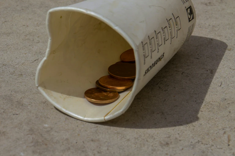 An old and empty Starbucks coffee cup lays on the floor with a few coins in it.