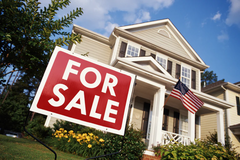 A low-angle shot of a house with a waving flag and a "for sale" sign.