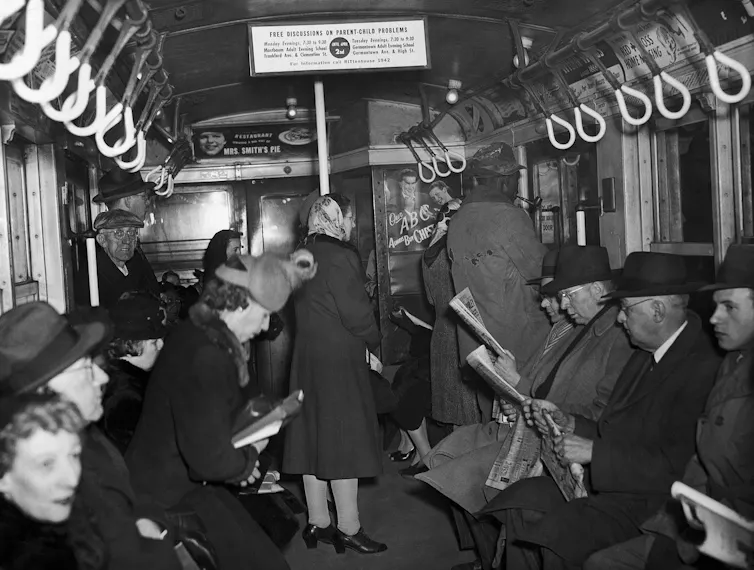 Black and white photo of men and women, some reading newspapers, in a subway car