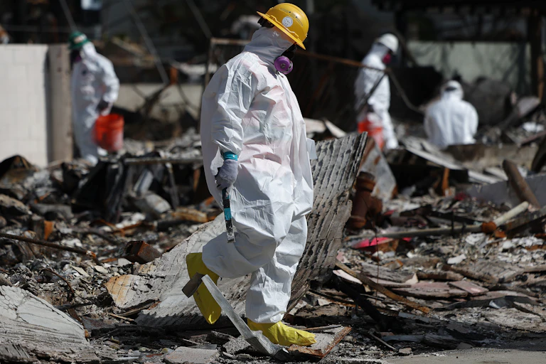 A person in a white protective suit, respirator and hard hat walks through an area filled with debris as other similarly clad people work in the background.