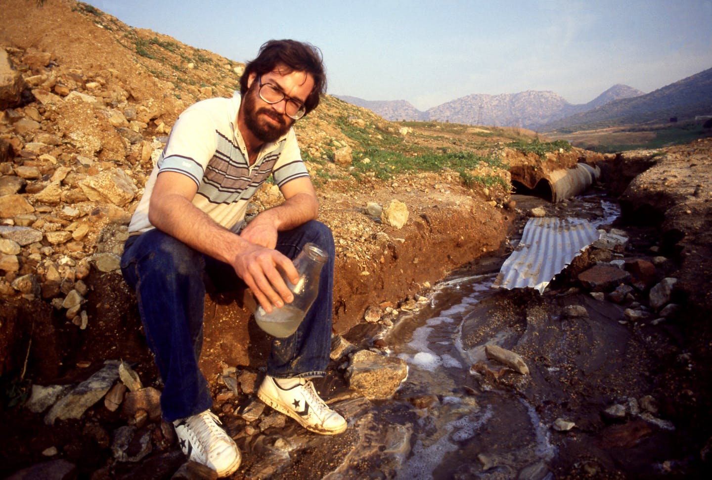 A person holds a clear jar of liquid while sitting on the ground in an area covered by rocks and dirt.