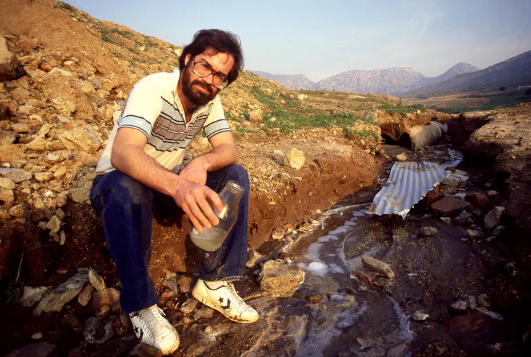 A person holds a clear jar of liquid while sitting on the ground in an area covered by rocks and dirt.