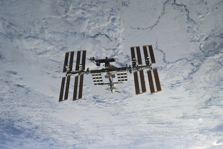 A ISS, que se parece com um cilindro com quatro braços de painéis solares, é mostrada flutuando no espaço, tendo como pano de fundo as nuvens da atmosfera terrestre.