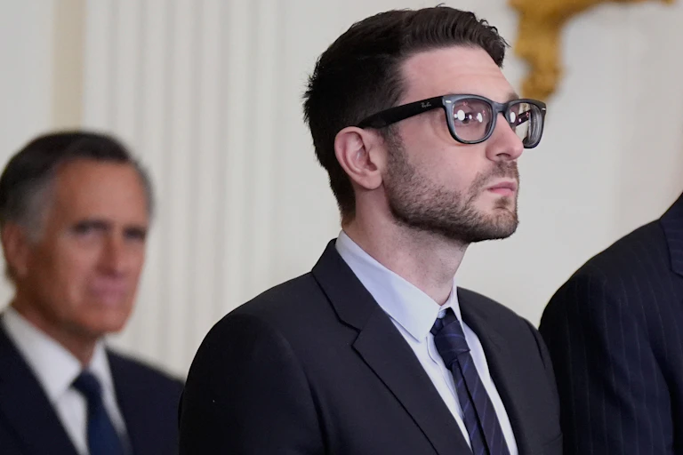 Man in a suit and tie and very dark-rimmed glasses looks out into the distance.