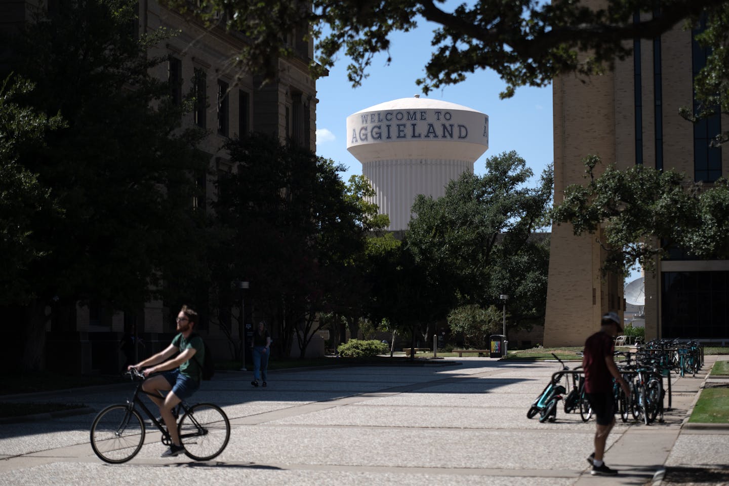 A large grassy and concrete space is seen with a water tower behind it and a person riding their bike, while another one walks.
