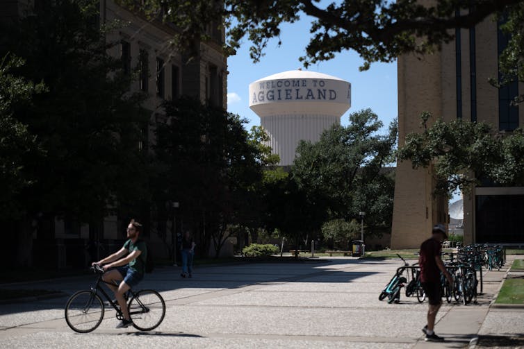 A large grassy and concrete space is seen with a water tower behind it and a person riding their bike, while another one walks.