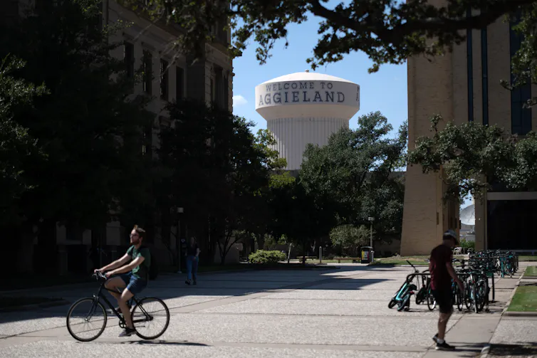 A large grassy and concrete space is seen with a water tower behind it and a person riding their bike, while another one walks.