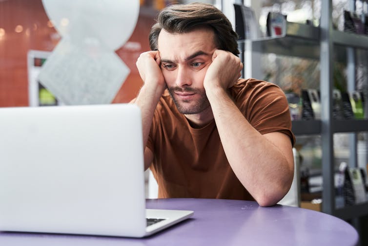A frustrated man stares at his laptop. He cradles his head in his fists.