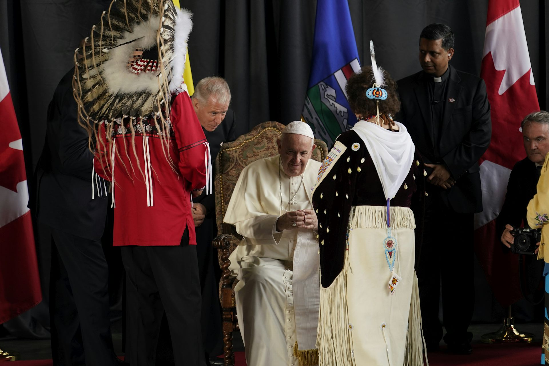 El hombre con vestidos blancos y la calavera blanca está sentada rodeada de mujeres con espacio blanco y plumas blancas y un hombre y un hombre con una pluma en la cabeza.