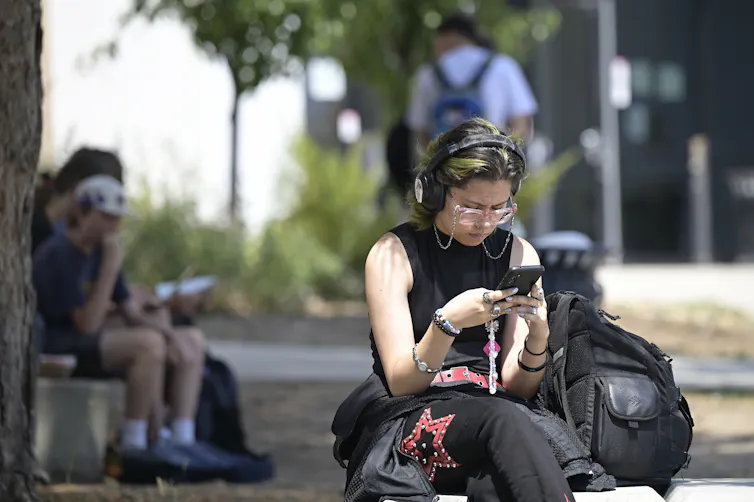 A white teen sits outside absorbed in her phone. She is wearing black clothing, glasses and headphones.