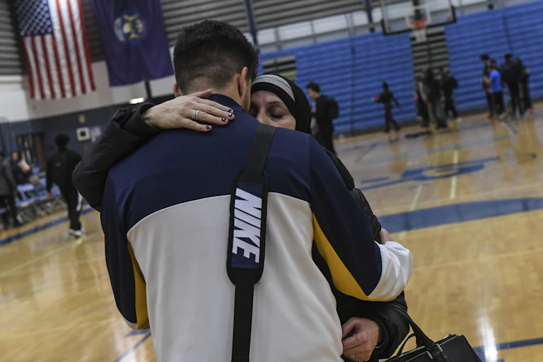 A tall young man wearing NIKE bag is hugged by a woman in hijab.