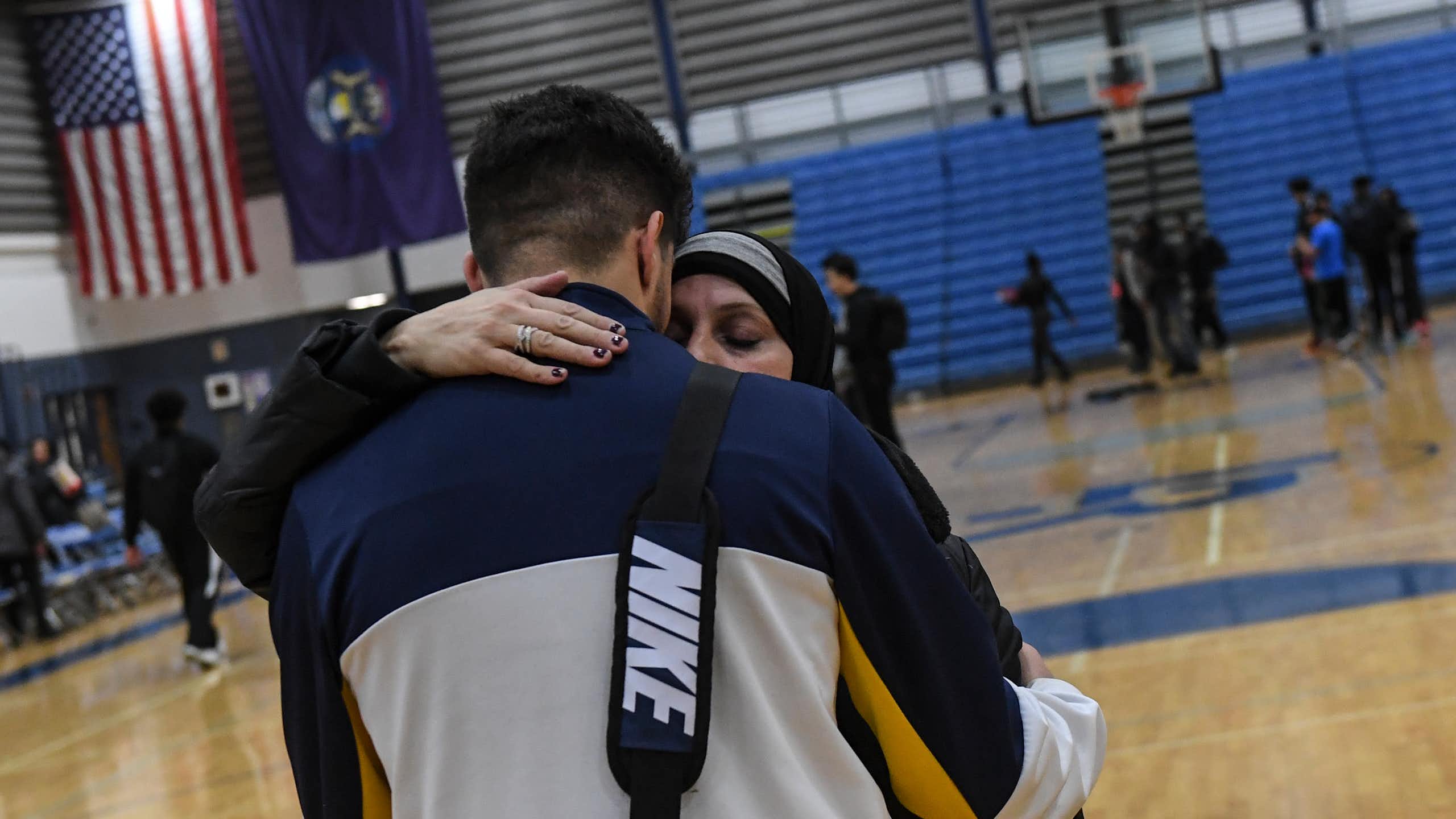 A tall young man wearing NIKE bag is hugged by a woman in hijab.