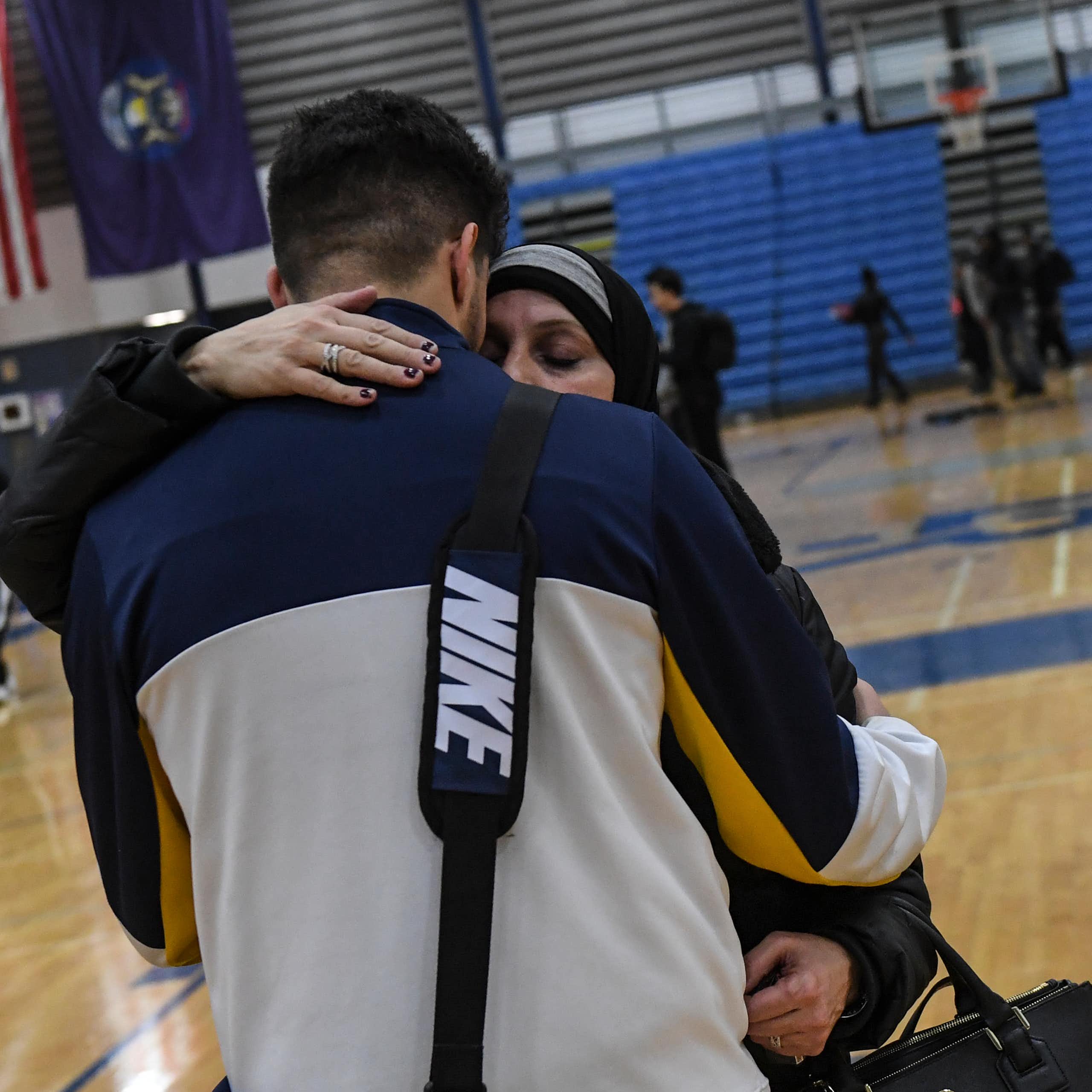 A tall young man wearing NIKE bag is hugged by a woman in hijab.