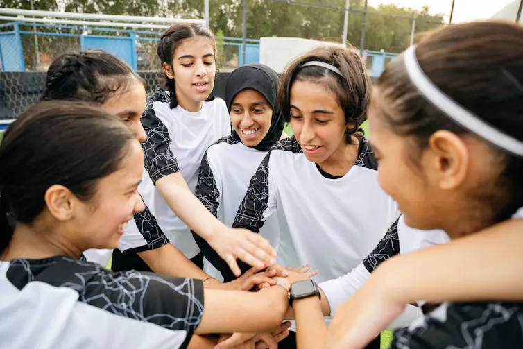 A group of tween girls put hands together in the middle of a circle. They are dressed in what look like soccer jerseys. Several are smiling