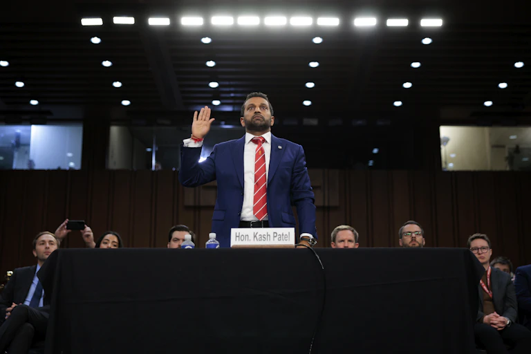 A man in a suit stands at a witness table, right hand raised.