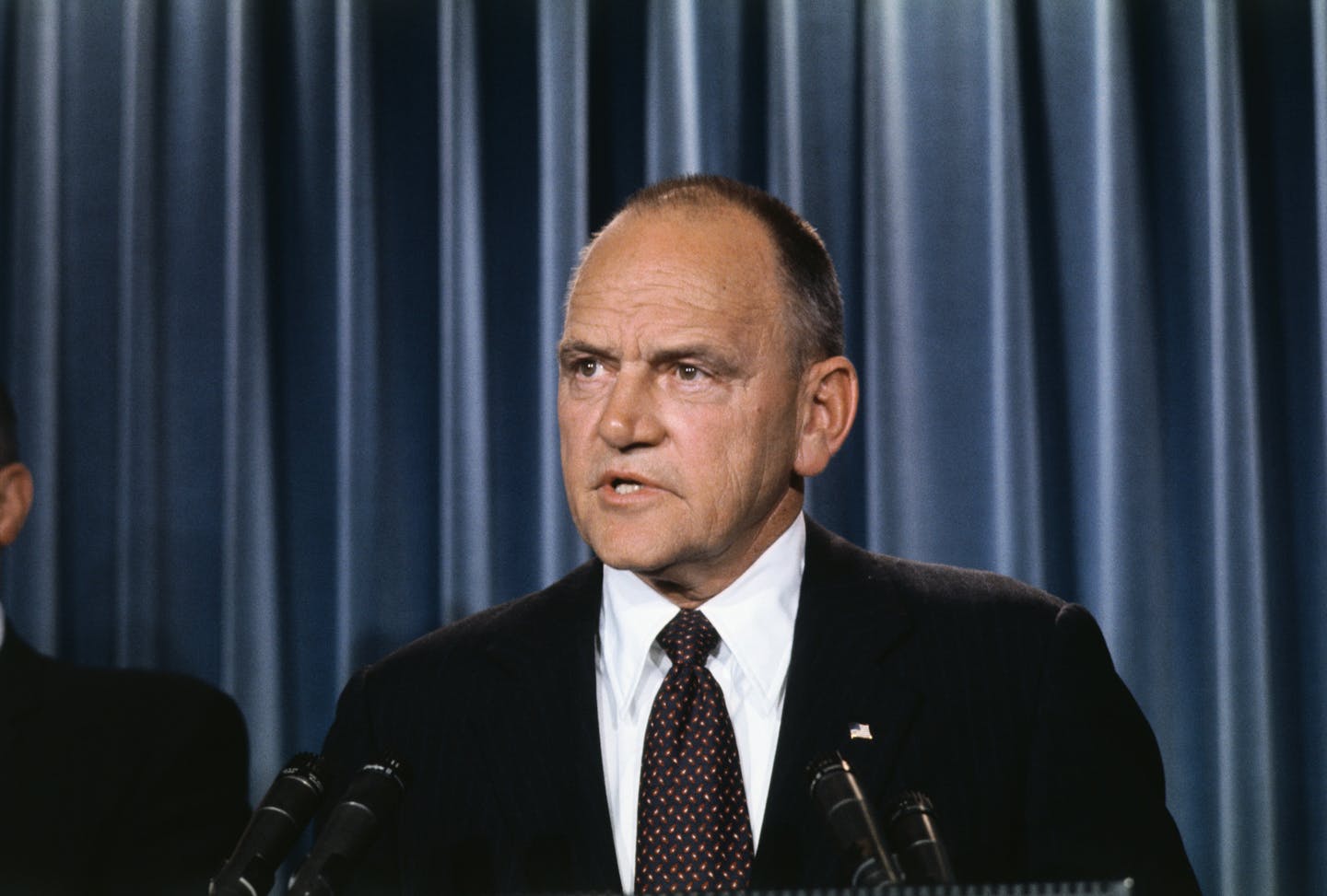 A man in a suit answers questions at a microphone.