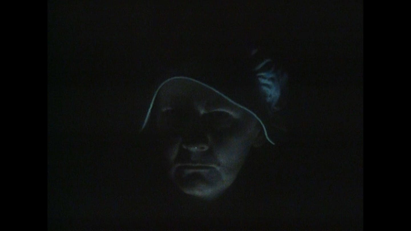 An old woman with a white hood peers sternly into the camera. It is dark so we only see a faint outline of her face.