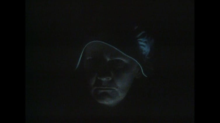 An old woman with a white hood peers sternly into the camera. It is dark so we only see a faint outline of her face.