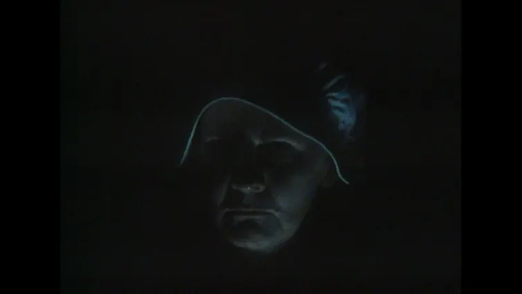 An old woman with a white hood peers sternly into the camera. It is dark so we only see a faint outline of her face.