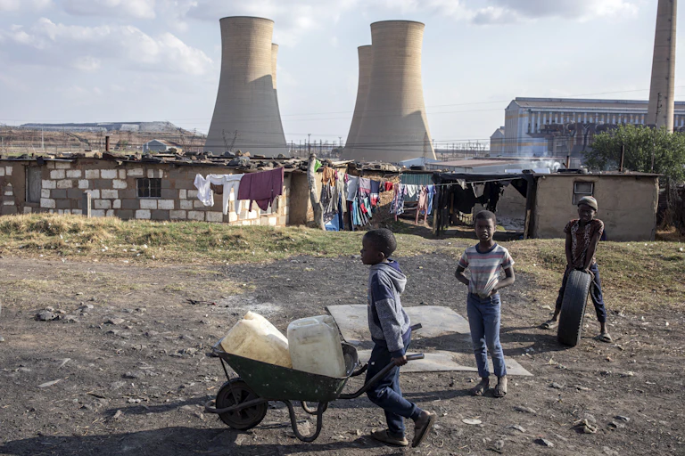 A coal fired power station with enormous towers and tin shacks built in front. A small boy wheels containers of water in a wheelbarrow.