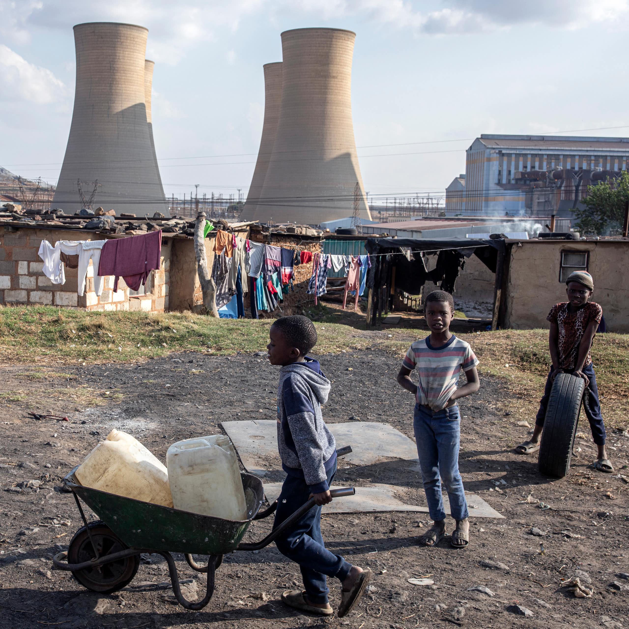 A coal fired power station with enormous towers and tin shacks built in front. A small boy wheels containers of water in a wheelbarrow.