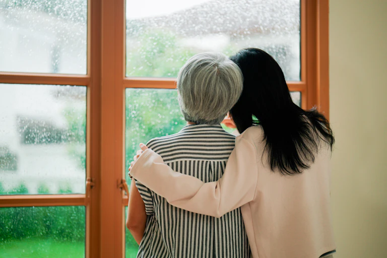 Younger woman embraces older woman as they look out a rainy window.