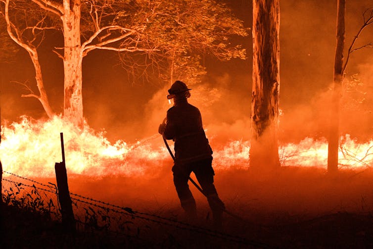 silhouette of firefighter spraying water on large bushfire.