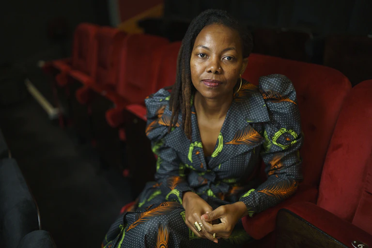 A young dreadlocked African woman in a wax print fabric dress sits in a red auditorium chair looking directly to camera, hands clasped.