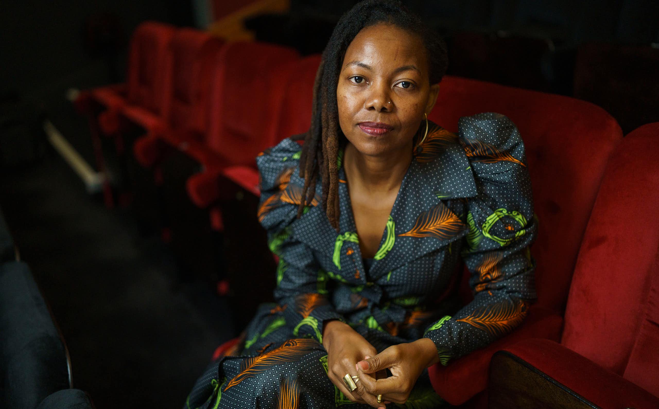 A young dreadlocked African woman in a wax print fabric dress sits in a red auditorium chair looking directly to camera, hands clasped.