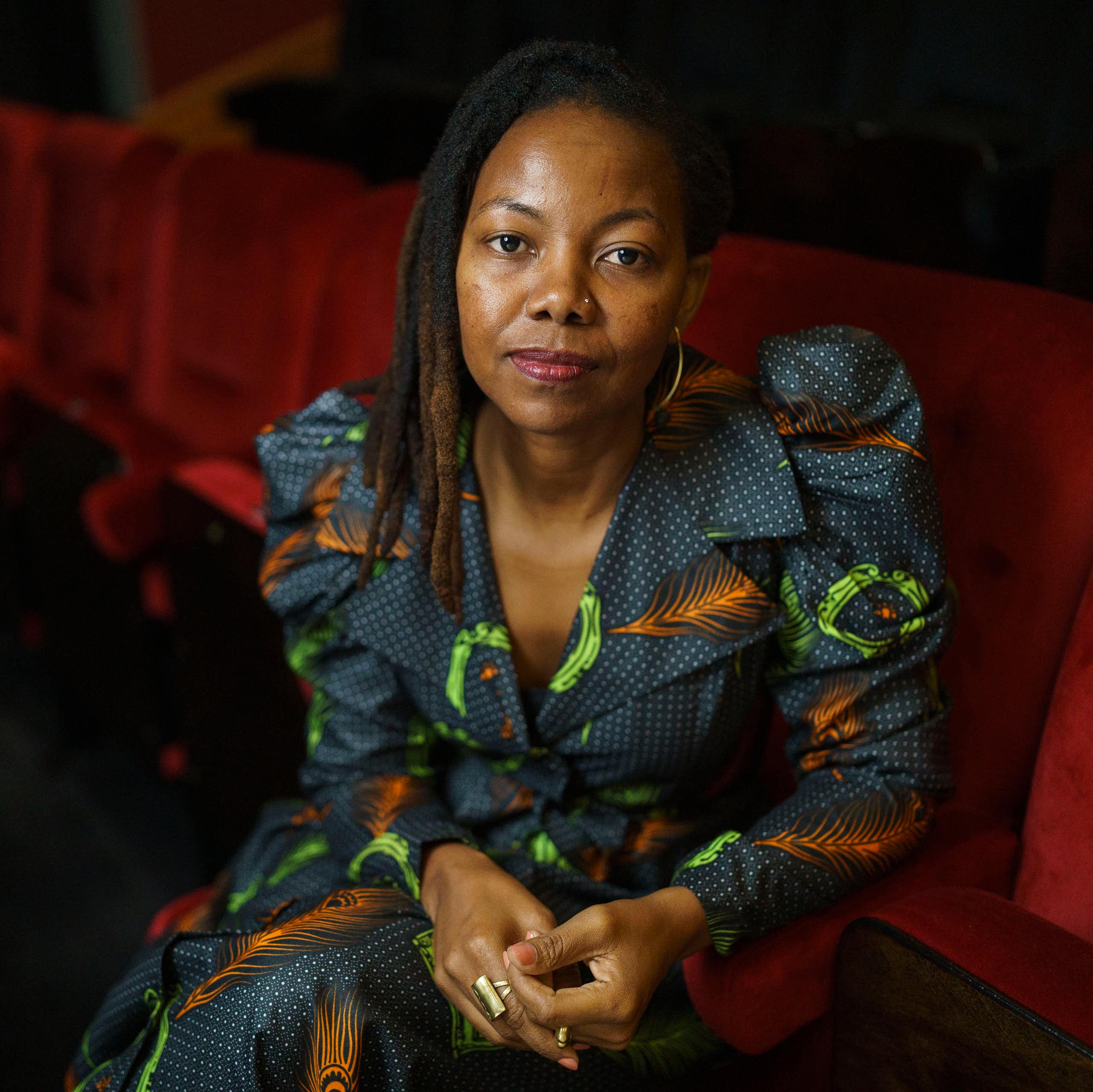 A young dreadlocked African woman in a wax print fabric dress sits in a red auditorium chair looking directly to camera, hands clasped.