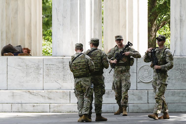 Los guardias nacionales armados se encuentran en un monumento con las personas sin hogar cercanas.