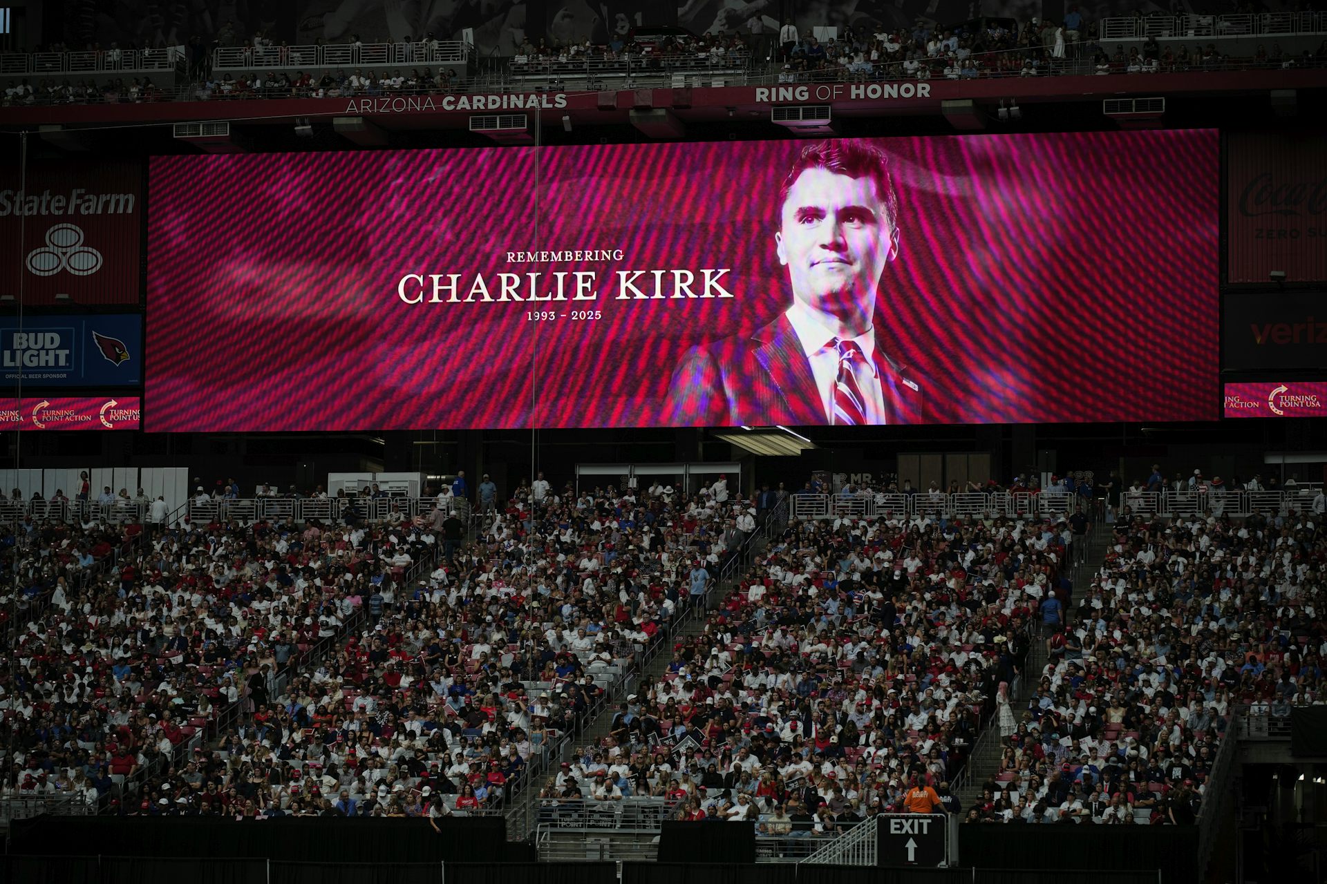 a man's face is projected on a screen behind a seated stadium crowd of hundreds