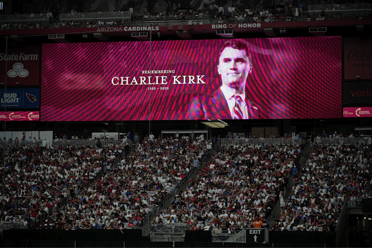 a man's face is projected on a screen behind a seated stadium crowd of hundreds