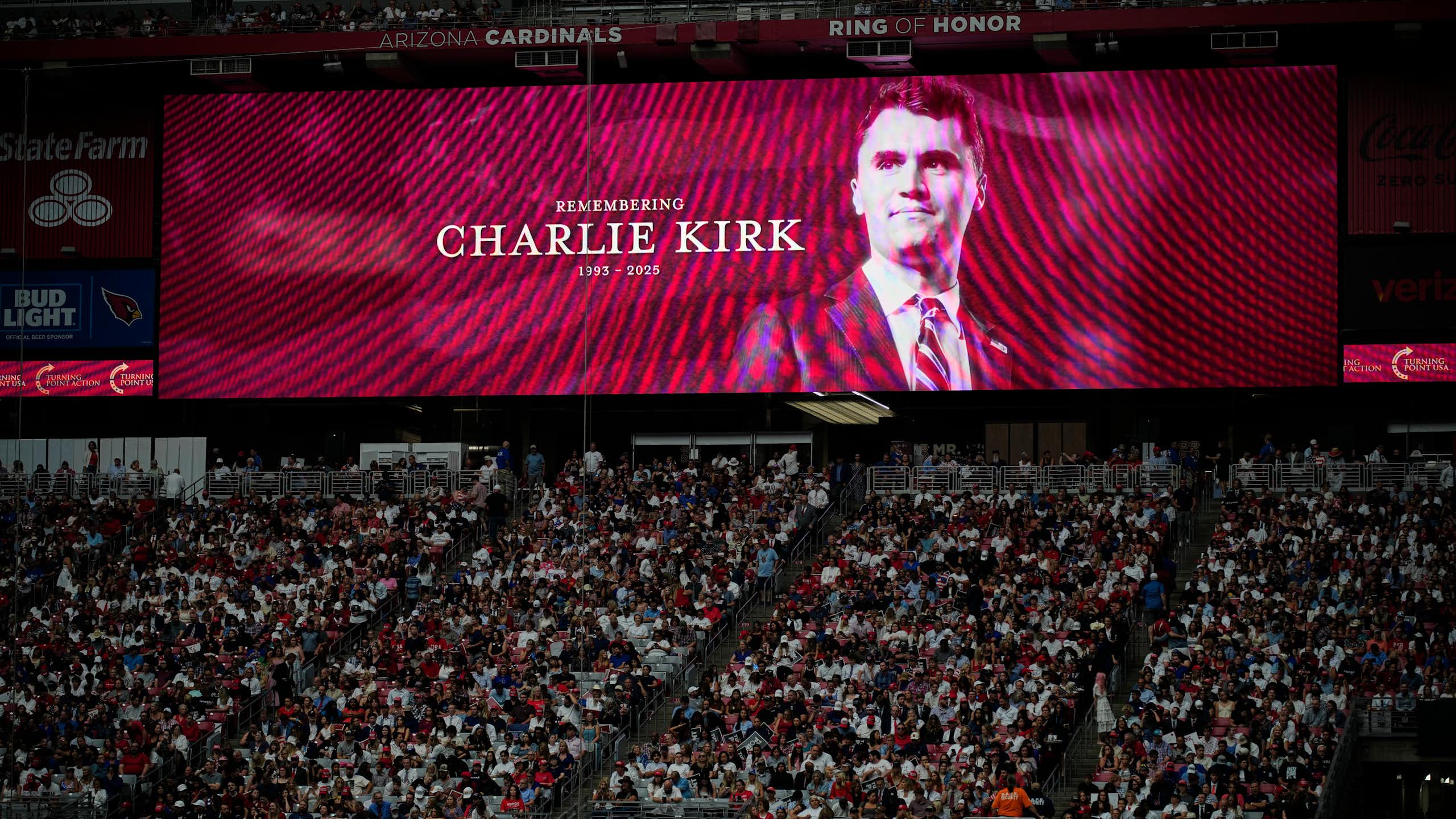 a man's face is projected on a screen behind a seated stadium crowd of hundreds