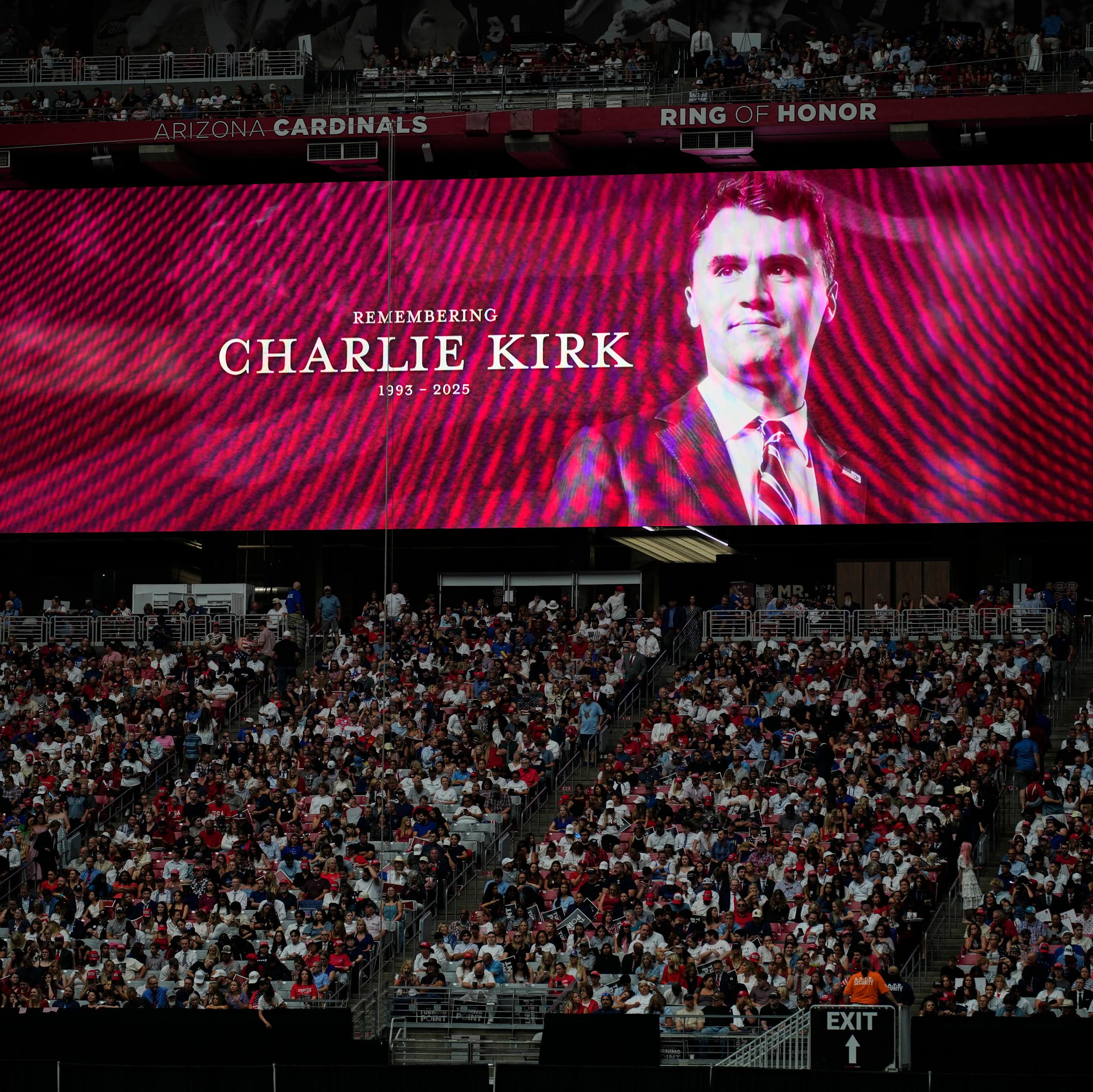 a man's face is projected on a screen behind a seated stadium crowd of hundreds