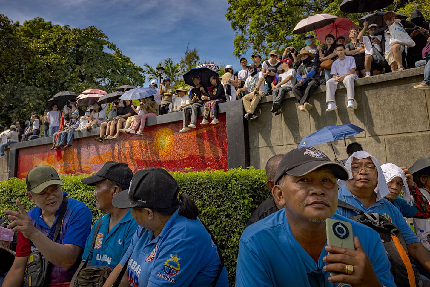 Rows of people sit on the ground and along a cement wall, all facing one way, under a blue sky.