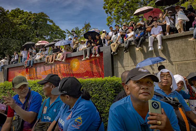 Rows of people sit on the ground and along a cement wall, all facing one way, under a blue sky.