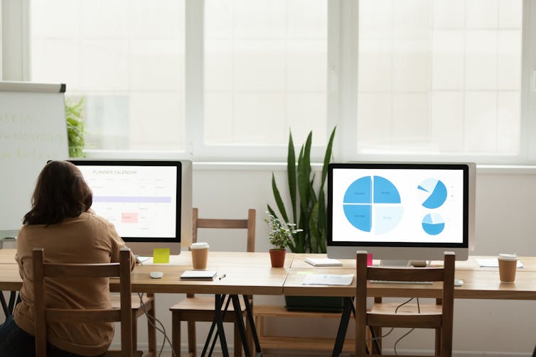 rear view person sat at computer in office, next to empty desk, graphics on screens