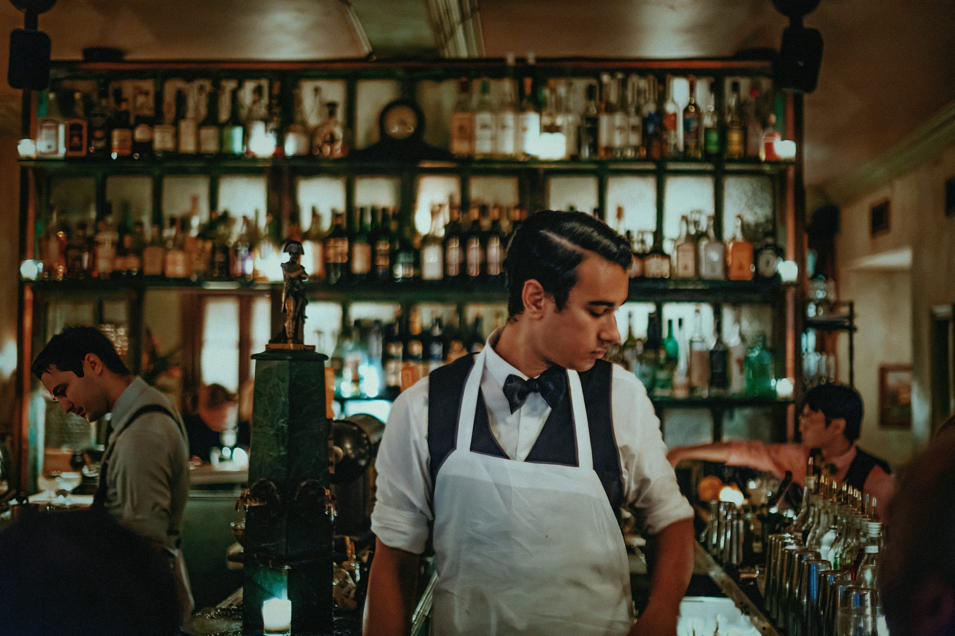 A bartender in an apron behind a counter. In the background, illuminated bottles of alcohol.
