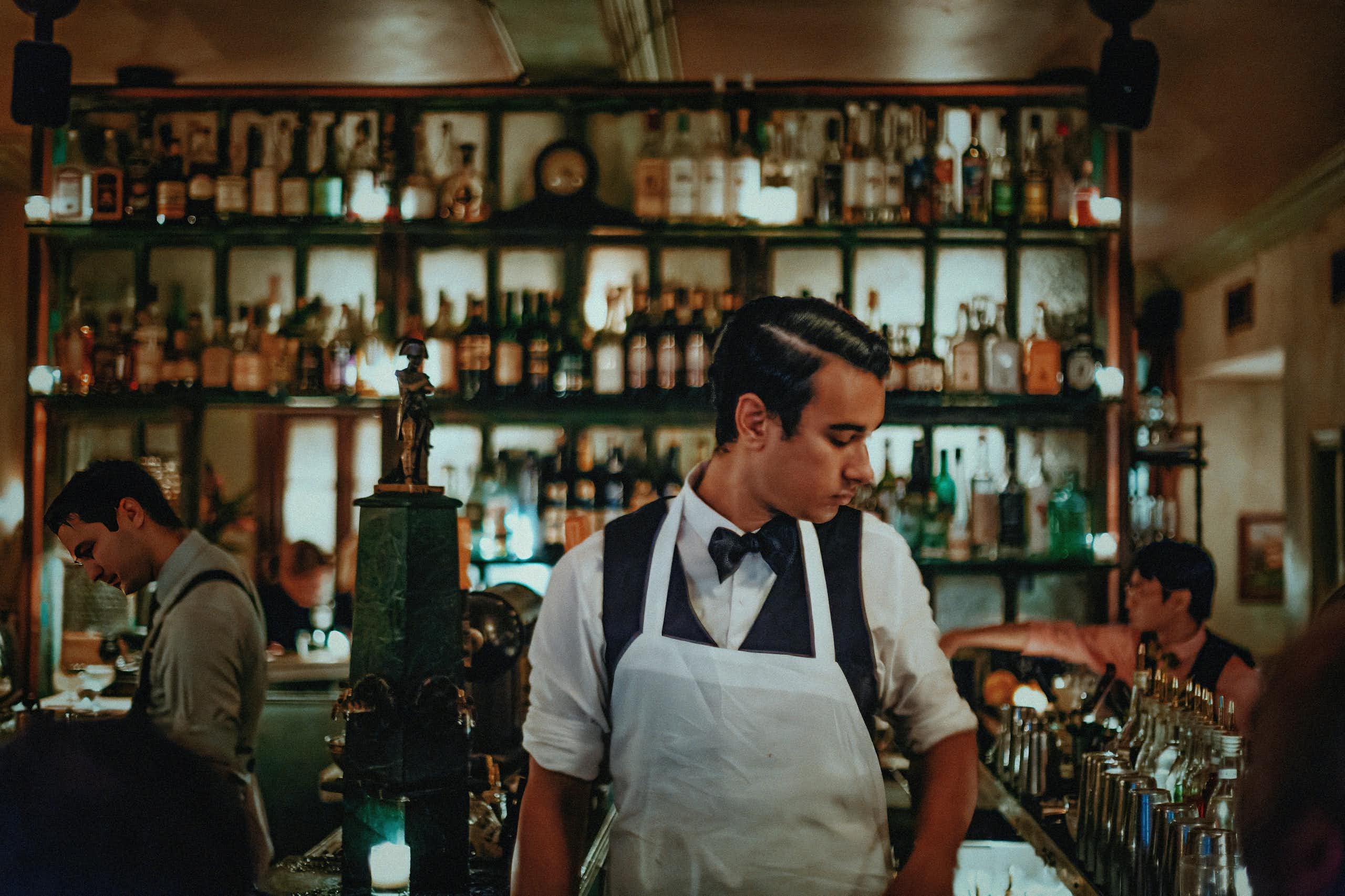 A bartender in an apron behind a counter. In the background, illuminated bottles of alcohol.