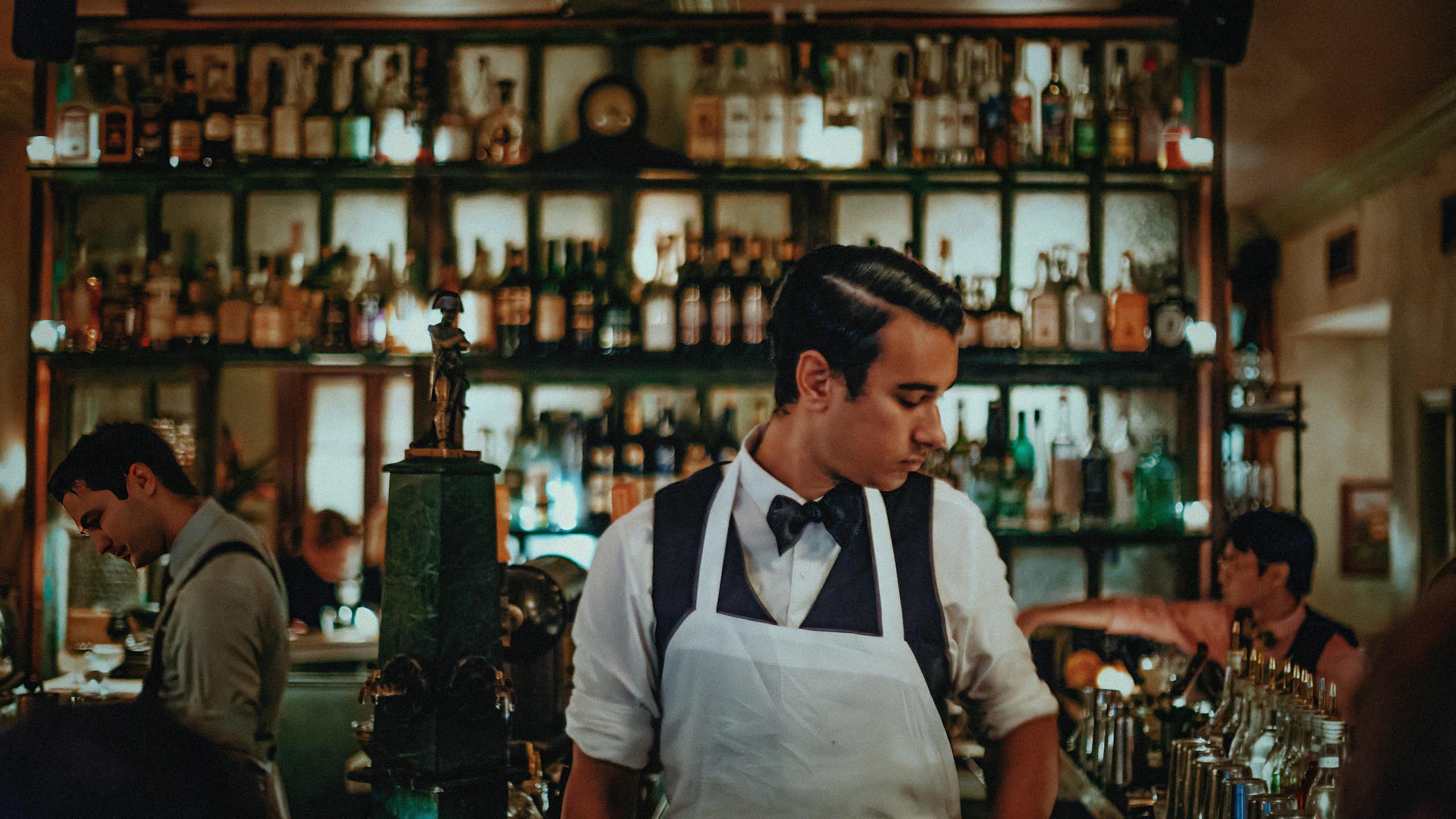 A bartender in an apron behind a counter. In the background, illuminated bottles of alcohol.