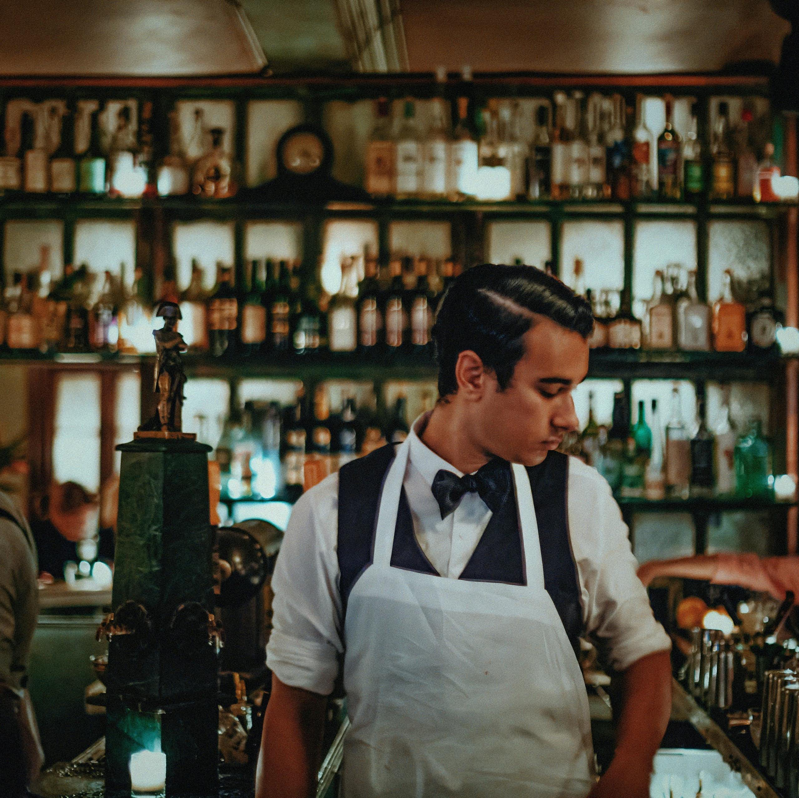 A bartender in an apron behind a counter. In the background, illuminated bottles of alcohol.