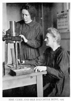 Maria Curie siedzi przy stole, na którym stoi sprzęt naukowy. Irena Curie stoi obok niej, bawiąc się sprzętem.