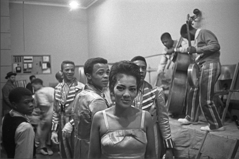 A black and white photo of a dance hall. A woman in a dress with shoulder straps looks directly into camera in front. Behind her are people dancing and a band on stage.