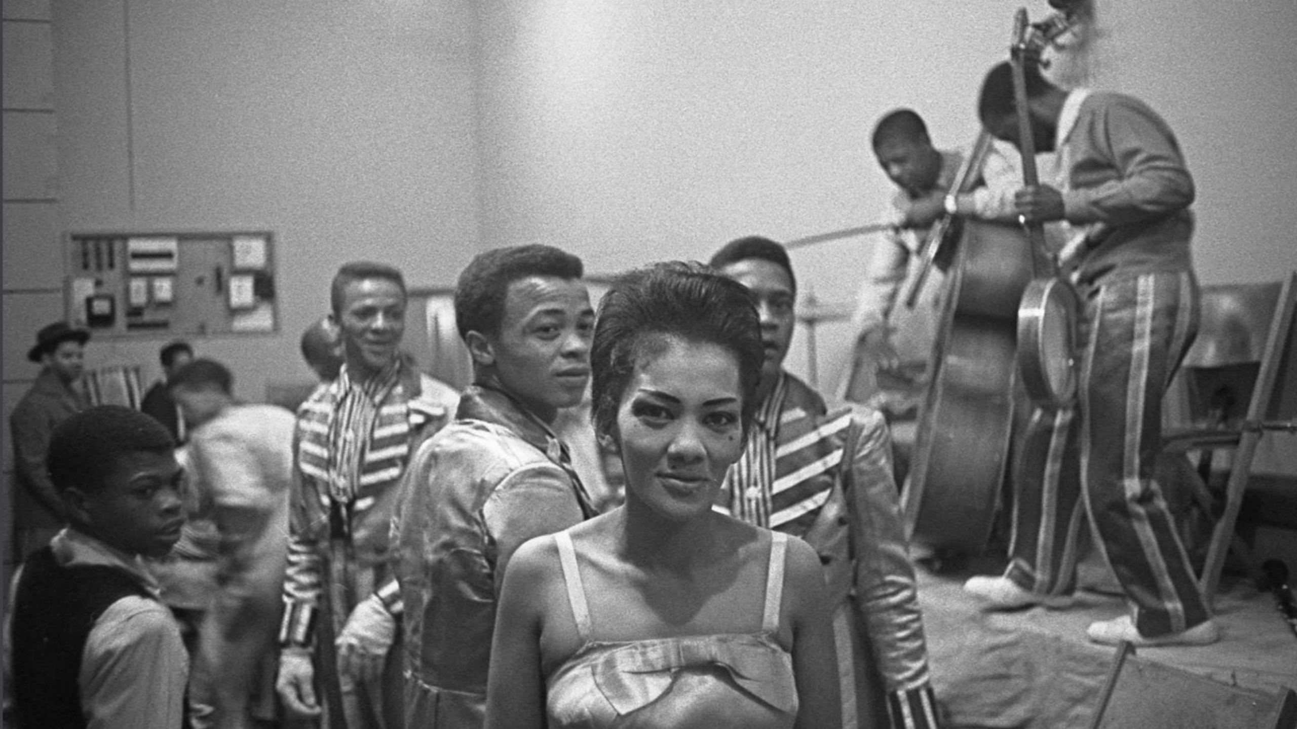 A black and white photo of a dance hall. A woman in a dress with shoulder straps looks directly into camera in front. Behind her are people dancing and a band on stage.